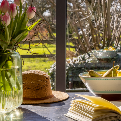 Dunowen fruits in a bowl, book hat and flowers on a table with an outside view.