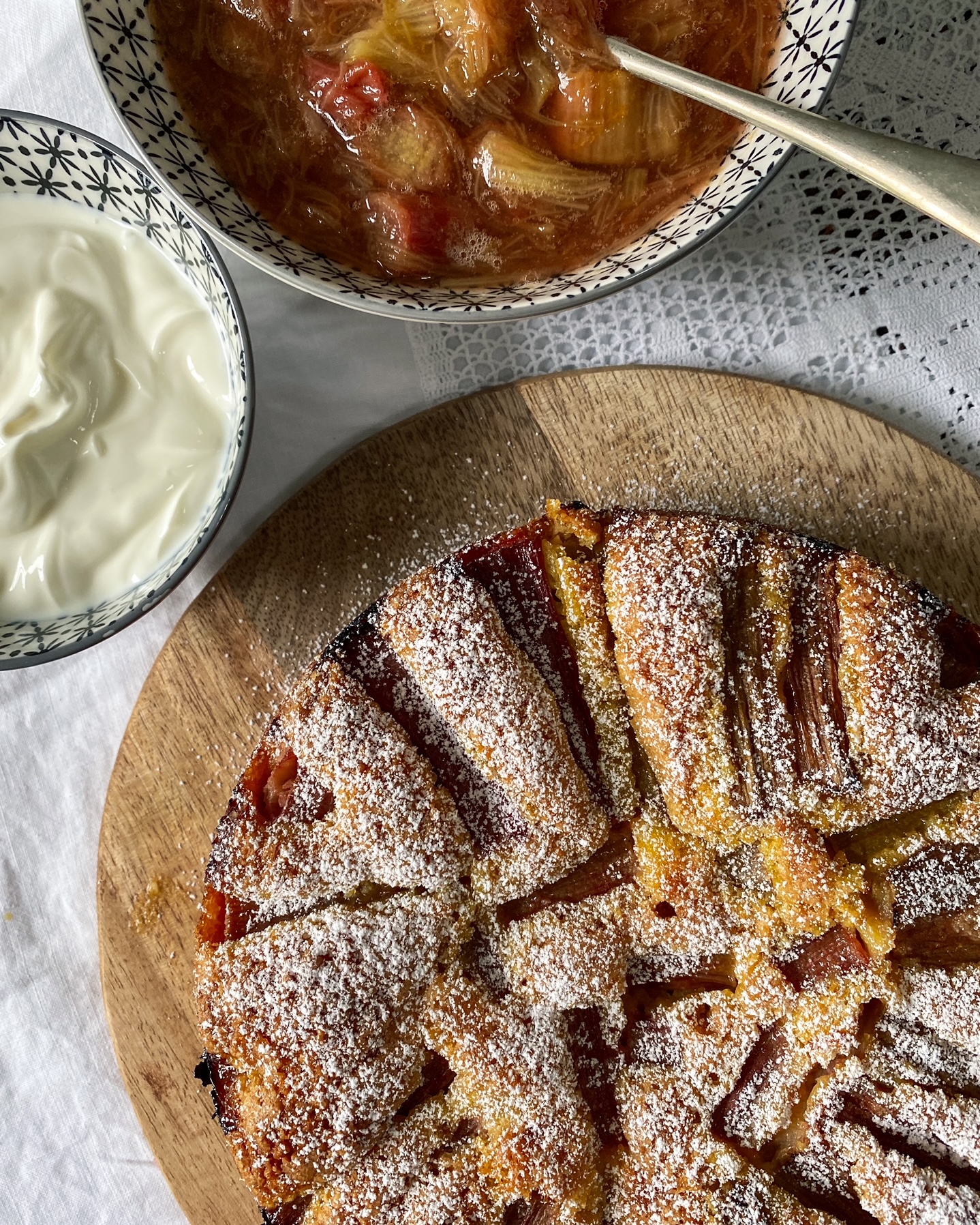 Rhubarb, Orange and Almond Cake on a wooden plate with glazing and whipped cream in bowls.