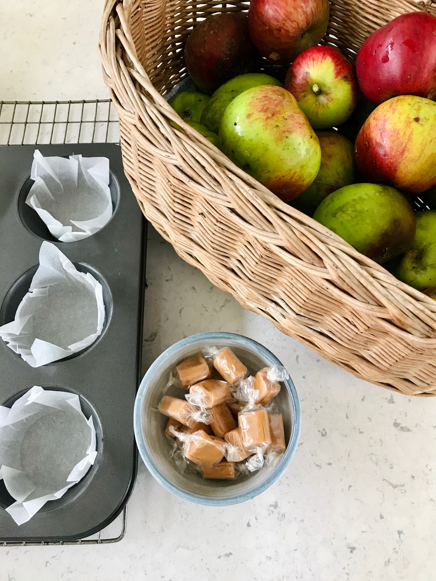 A muffin tin, apples in a basket and caramel candy in a bowl on a kitchen countertop.