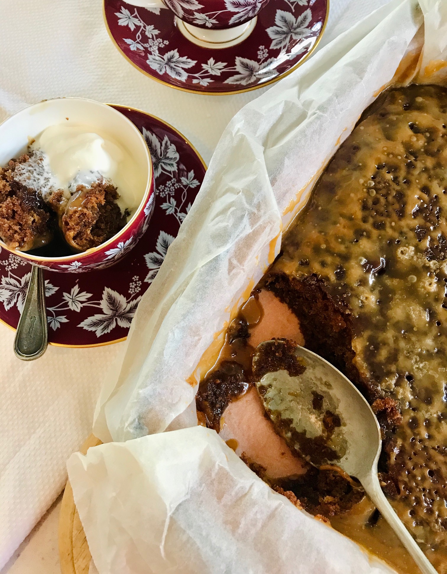 Sticky toffee pudding cake in a cake pan next to Sticky toffee pudding cake in a cup on a plate on a table.